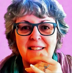 A picture of a woman's head against a pink background. She has blue highlights in her hair and dark blue glasses. She is staring straight at you, with a smile and her left hand supporting her chin. 