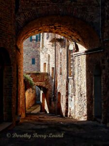 This is a photograph looking from shade under an archway to sunshine beyond. The shade is very dark in the foreground under the archway which is part of a medieval town wall in Italy. Looking through the arch there is brilliant, bright sunshine highlighting the buildings behond, and you can see the traditional stone built architecture, windows and shutters. In the middle distance is another smaller archway that connects one side of the street with the other.